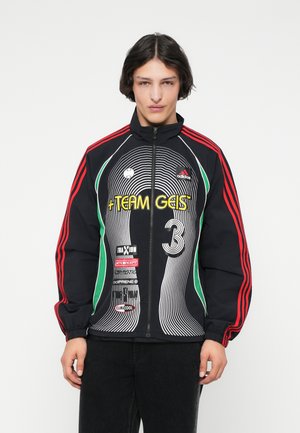 Joven con una chaqueta negra de Adidas con cremallera y rayas rojas, logos multicolores y el texto "TEAM GEIST 3", de pie frente a un fondo blanco.