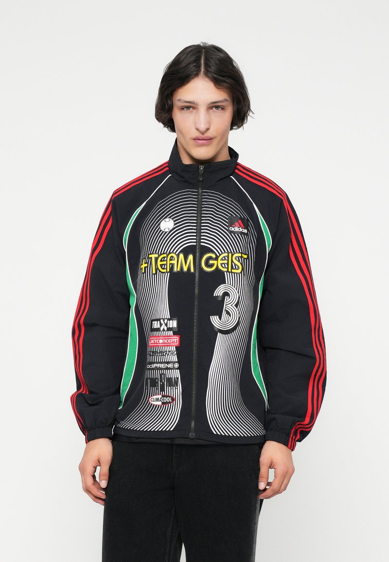 Ungdom som har på seg en svart Adidas-jakke med glidelås og røde striper, flerfargede logoer og teksten "TEAM GEIST 3", stående mot en hvit bakgrunn.