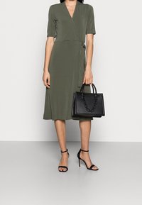 Khaki-grön omvikningsklänning med korta ärmar, en slejf i midjan och en svängig kjol, kombinerad med en svart handväska med kedjedetalj och klackskor med ankelsöm.