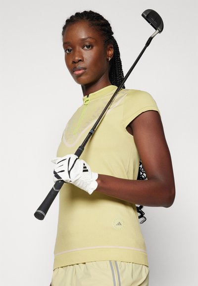 Chemise de golf jaune clair à manches courtes avec un col haut, fermeture éclair verte et détails de logo. Gant de golf blanc tenant un club noir.