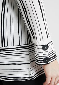 Zwart-wit gestreepte pullover met lange mouwen, voorzien van een omgeslagen boord en drie decoratieve zwarte knopen bij de mouwzoom.