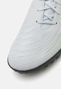 Hellgraue Fußballschuhe mit einem strukturierten Obermaterial, das Wellenmuster und reflektierende Akzente aufweist, schwarze Gummisohle und in das Design integrierte Schnürsenkel.