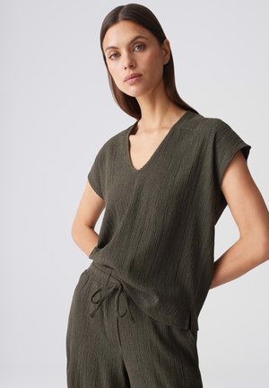 Donna con capelli lisci e scuri che indossa un top verde oliva testurizzato con scollo a V e pantaloni abbinati con coulisse, su uno sfondo grigio uniforme.