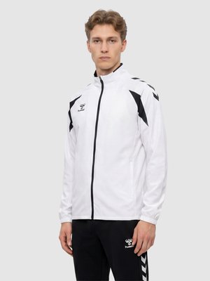 Junger Mann trägt eine weiße Sportjacke mit Reißverschluss und schwarzen Chevron-Akzenten sowie schwarze Hose mit passendem Logo und Chevron-Streifen.