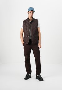 Καπιτονέ καφέ γιλέκο με κουμπιά snap, φορεμένο πάνω από ένα λευκό πουκάμισο. Σκούρα καφέ παντελόνια και μαύρα εσπαντρίγια, συμπληρωμένα από ένα ανοιχτό μπλε καπέλο.