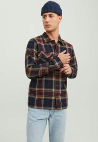 Camisa de franela a cuadros en azul marino, burdeos y beige. Presenta dos bolsillos en el pecho y cierre con botones en la parte delantera. Llevada con jeans de color azul claro y un gorro de punto.