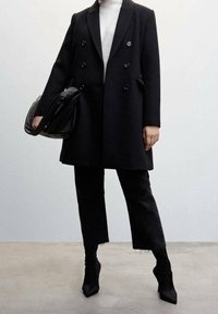 Persoon in een zwart dubbelzijdig jasje, zwarte cropped broek, zwarte haklaarzen, witte coltrui, en met een grote zwarte handtas in de hand.