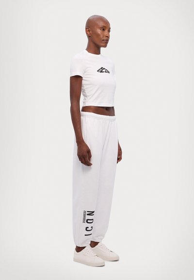 T-shirt blanc court avec design de logo noir, associé à un pantalon de survêtement blanc avec impression de logo sur le côté et poignets élastiques. Baskets blanches.