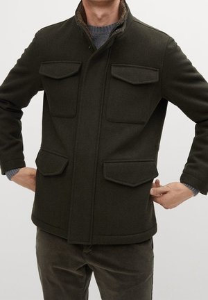 Mężczyzna w ciemnozielonej kurtce z czterema kieszeniami z klapkami i szarym swetrze pod spodem, z rękami opartymi na biodrach.