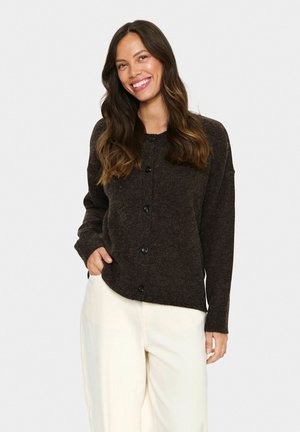 Young woman smiling, wearing a dark button-up cardigan and light-colored wide-leg trousers, standing with one hand in pocket.