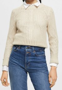 Beige gestrickter Pullover mit einer strukturierten offenen Maschenweise, Rundhalsausschnitt und gerippten Bündchen, kombiniert mit hochgeschnittenen Jeans.