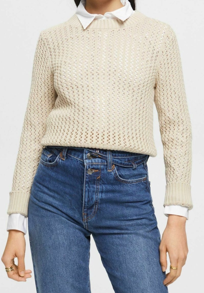 Beige gestrickter Pullover mit einer strukturierten offenen Maschenweise, Rundhalsausschnitt und gerippten Bündchen, kombiniert mit hochgeschnittenen Jeans.