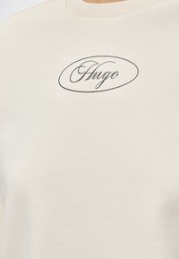 Pull couleur crème avec « Hugo » écrit en noir cursif à l’intérieur d’un ovale fin sur le haut de la poitrine.