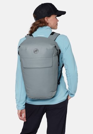 Persoon die een lichtblauwe fleece, zwarte pet en een grote grijze rugzak met Mammut-logo draagt, staand met één hand in de zak, zijwaarts gekeerd.