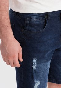 Pantalones cortos de mezclilla azul oscuro con un patrón desgastado, con un desgarro visible y un ligero desvanecimiento en la tela. Presenta bolsillos frontales.