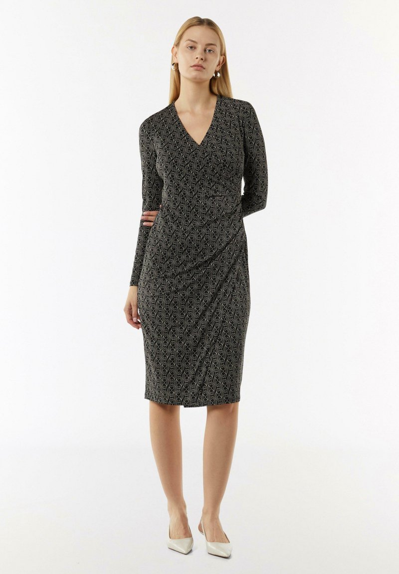 Schwarzes Kleid mit langen Ärmeln, V-Ausschnitt und einem strukturierten Muster. Figurbetonte Form, knielanger Saum, kombiniert mit weißen spitzen Schuhen.