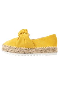 Espadrille de ante amarillo con suela de yute trenzado grueso, suela de goma blanca con bordes en forma de dientes y un gran lazo anudado en la parte superior.