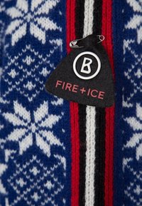 Étiquette triangulaire noire avec le logo "B" et le texte "FIRE + ICE" épinglée sur un tissu tricoté bleu et blanc à motif de flocons de neige avec des rayures rouges et noires.