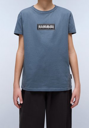 Osoba nosząca przygaszony niebieski t-shirt z krótkim rękawem, z czarno-białym prostokątnym logo na środku klatki piersiowej i czarne spodnie.