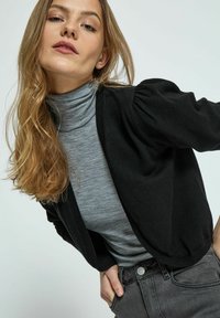 Schwarze Cardigan mit Puffärmeln über einem grauen Rollkragenpullover, kombiniert mit grauen Jeans. Der Cardigan hat gerippte Kanten und eine entspannte Passform.