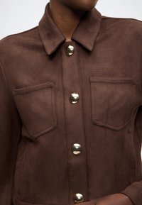 Braune Wildlederjacke mit einem Kragen, glänzenden goldenen Knöpfen und zwei Brusttaschen. Weiche Textur mit einem sauberen und modernen Design.