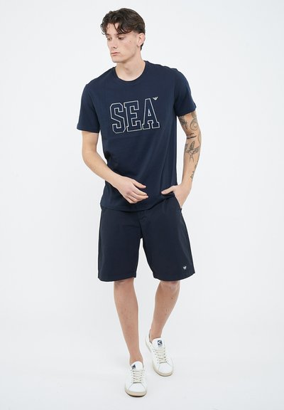 Giovane uomo con tatuaggi, indossa una maglietta blu navy con la scritta "SEA", pantaloncini abbinati e scarpe da ginnastica bianche, in piedi con una mano in tasca su sfondo bianco.