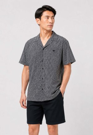 Uomo che indossa una camicia a maniche corte nera e bianca con bottoni e pantaloncini neri, in piedi con una mano in tasca.