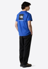 Blå bomulds T-shirt med korte ærmer, prydet med et sort firkantet grafisk print og hvid tekst på ryggen. Sammen med sorte bukser.