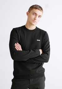 Črna olemka s kratkimi rokavi iz mehkega blaga, ki ima na prsih majhen bel logotip "BOSS". Dolgi rokavi z rebrastimi manšetami.