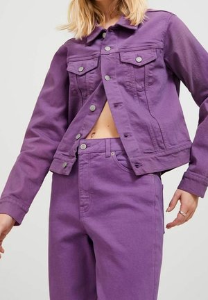 Kvinde iført en opknappet lilla denimjakke og matchende lilla højtaljede jeans, der viser en smule af maven mod en ensfarvet baggrund.