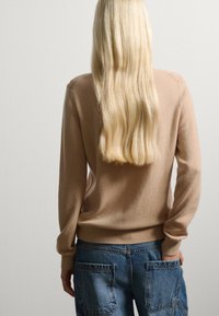 Maglione beige a maglia con maniche lunghe, scollatura rotonda e polsini a coste, abbinato a pantaloni in denim blu con grandi tasche posteriori.