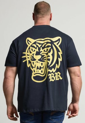 Hombre de espaldas con camiseta azul marino con un gran gráfico amarillo de una cara de tigre rugiente y las letras "BR" en la espalda.