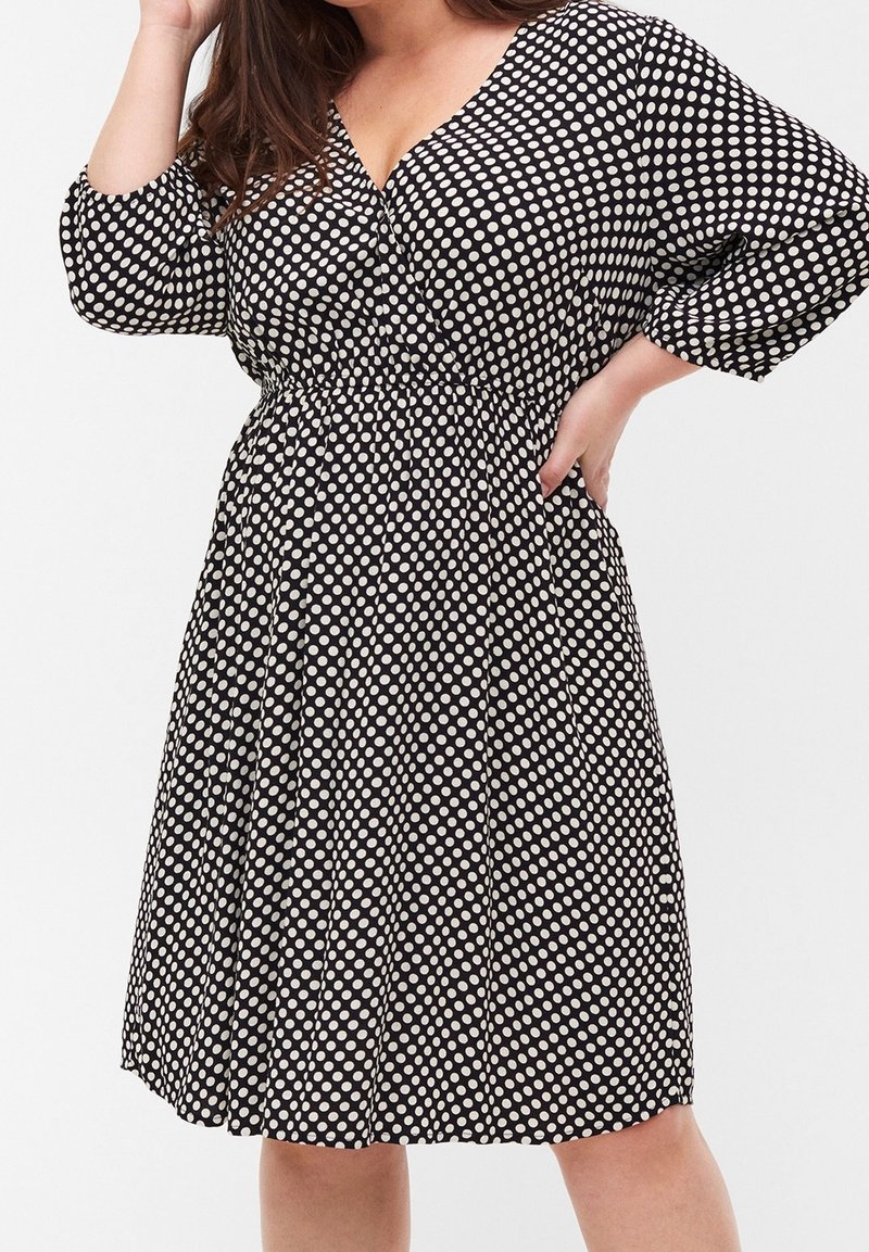 Femme portant une robe noire longueur genou avec des pois blancs, manches trois-quarts et une taille froncée, posant avec une main sur la hanche.