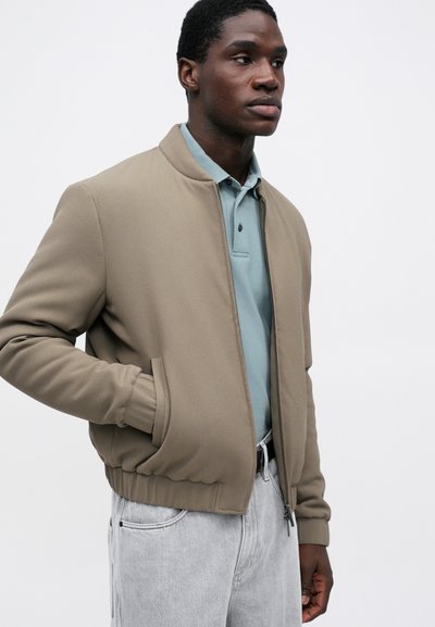 Bomberjakke i lys brun teksturert stoff, med glidelås foran, ribbestrikkede mansjetter og linning, kombinert med en lys blå polo og grå bukser.