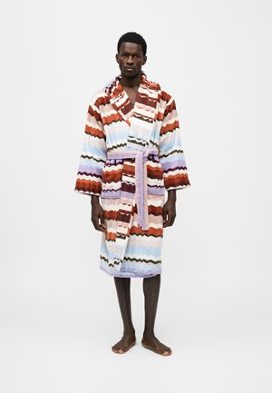 Un homme se tient pieds nus sur un fond uni et présente une robe de chambre ceinturée à rayures en zigzag colorées.