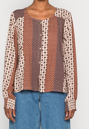 Kvinde iført en knappet bluse med lange ærmer med lodrette brune og beige geometriske mønstre og blå denimbukser.