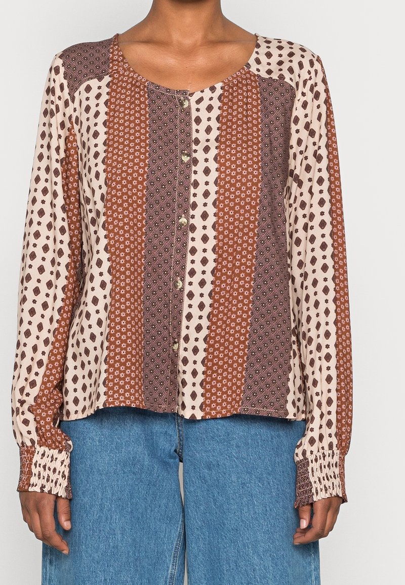 Femme portant un chemisier à manches longues boutonné avec des motifs géométriques verticaux marron et beige et un pantalon en jean bleu.