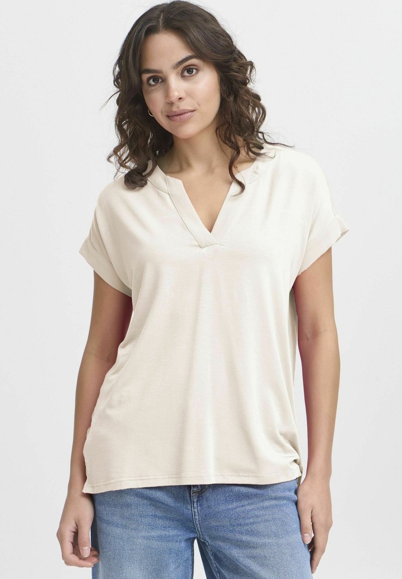Top beige a maniche corte con scollo a V e risvolti, realizzato in tessuto morbido, caratterizzato da una vestibilità ampia e una texture liscia. Indossato con jeans blu.