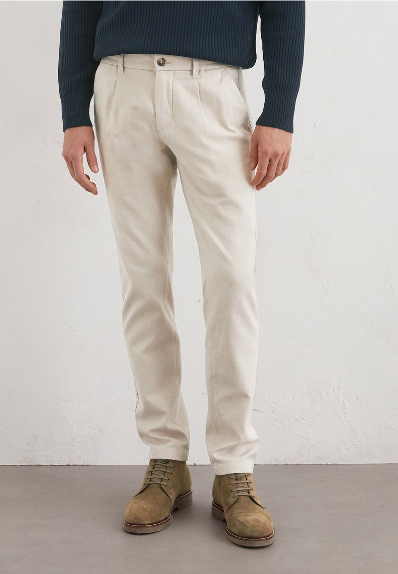 Homme portant un pantalon beige, des bottes lacées vert olive et un pull côtelé bleu foncé à manches longues, debout sur un sol gris.