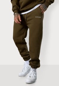 Olivgrüne Sweatpants mit elastischen Bündchen und einem Kordelzugbund, versehen mit einem weißen Logo auf dem linken Oberschenkel und kombiniert mit weißen Turnschuhen.