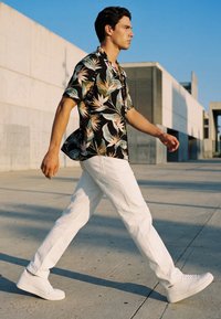 Young man walking outdoors wearing a black floral shirt, white pants, and white sneakers against a modern urban background.