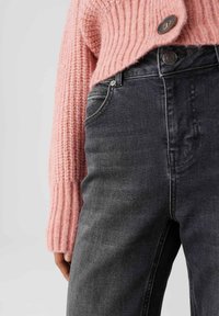 Ravvicinato di una persona che indossa jeans neri sbiaditi e un maglione rosa lavorato a maglia con grandi dettagli a bottoni.
