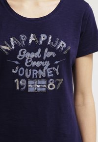 Camiseta de color azul marino con cuello redondo, que presenta el texto en lentejuelas plateadas "NAPAPIJRI Bueno para Cada Viaje 1987" y un diseño de bandera.