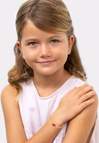 Bracelet jaune avec un charme de pastèque rouge, porté au poignet d'un enfant aux longs cheveux bruns et portant un haut sans manches rose clair.