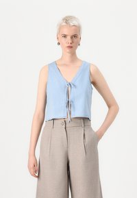 Lichtblauw cropped mouwloos topje met een v-hals en striksluiting aan de voorkant, gecombineerd met high-waisted, lichtbruine wijde broeken.