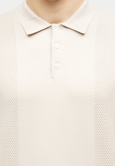 Μπεζ κοντομάνικο πόλο μπλουζάκι με τρία κουμπιά και υφασμάτινες πλεκτές πινελιές στις πλευρές, που φοριέται από ένα άτομο.