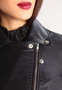 Svart läderjacka med silverfärgad dragkedja och tryckknappar, med en volangkrage av svart tyg och slät textur. Närbild av halsringningen och detaljer.