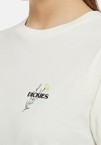 Kermanvärinen t-paita pehmeästä puuvillasta, jossa on kukkakuva ja musta-viher "Dickies" -logo vasemmassa rintaosassa.