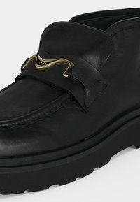 Μαύρο δερμάτινο loafer με πάχη ελαστική σόλα και χρυσή διακοσμητική μεταλλική αγκράφα στην κορυφή, με διακριτικές ραφές.