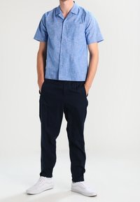 Man som bär en ljusblå kortärmad skjorta med knappar, mörkblå byxor och vita sneakers, står med händerna i fickorna mot en vit bakgrund.